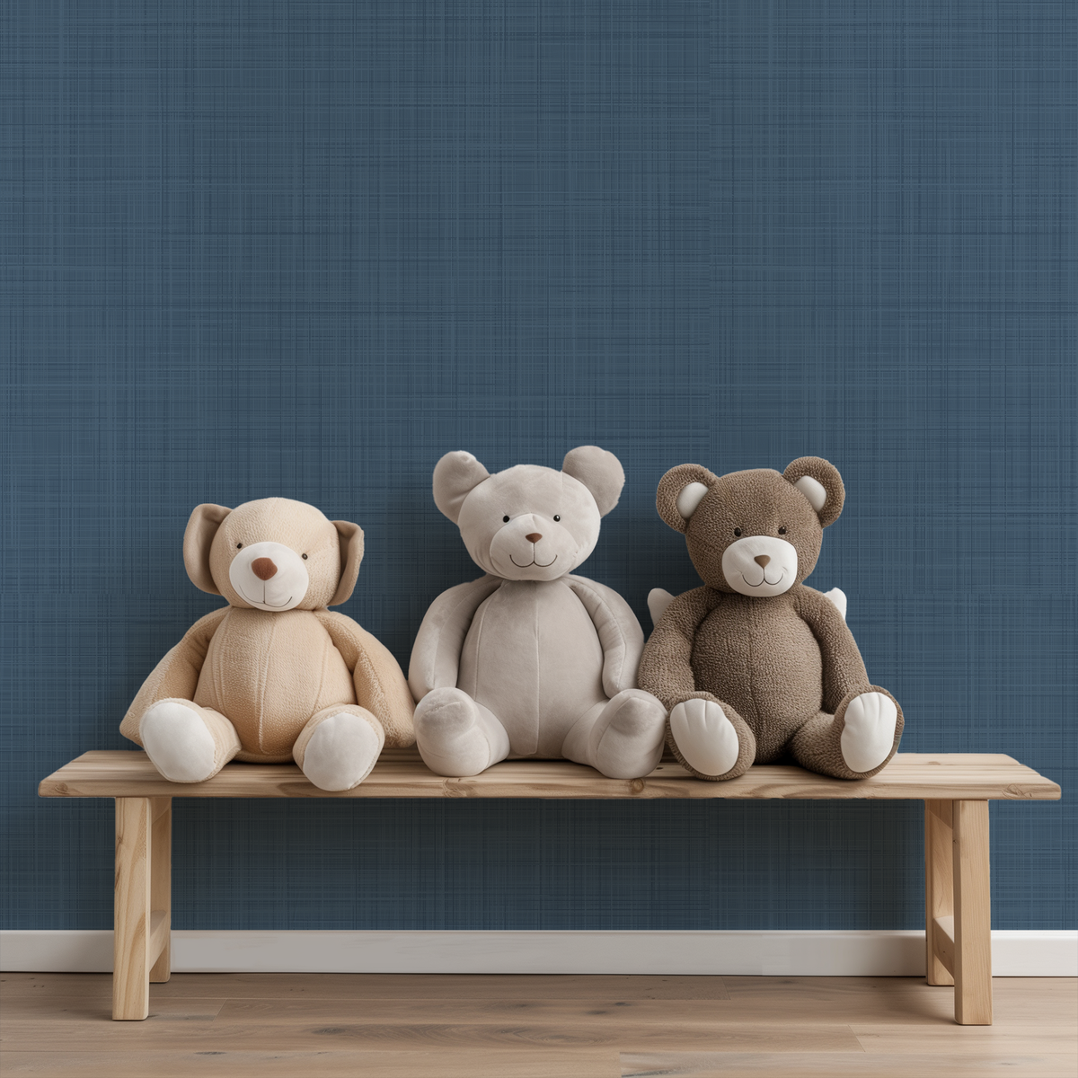 Three plush teddy bears in beige, light gray, and brown sit side by side on a light wood bench against a faux textured dark blue grasscloth wallpapered wall