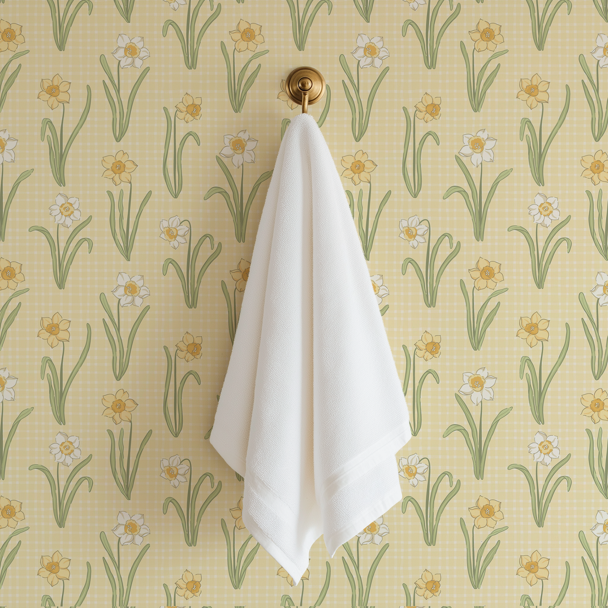 A white cotton hand towel hangs from a round gold wall hook against a yellow wallpaper patterned with white daffodils and green stems.