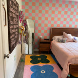 Check wallpaper (orange, pink, and light blue checked) in girl's bedroom with queen bed, pink quilt. Big, bright floral rug under bed.