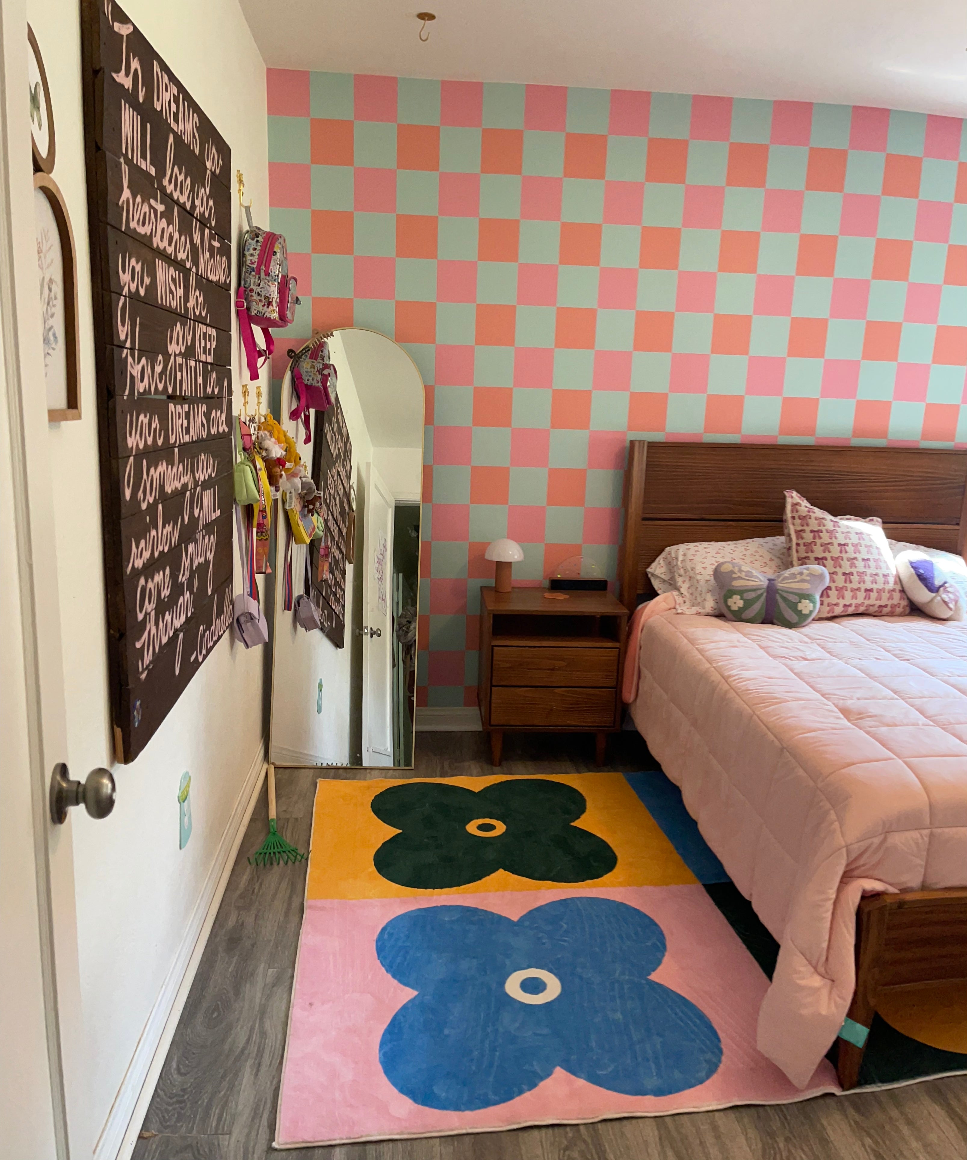 Check wallpaper (orange, pink, and light blue checked) in girl's bedroom with queen bed, pink quilt. Big, bright floral rug under bed.