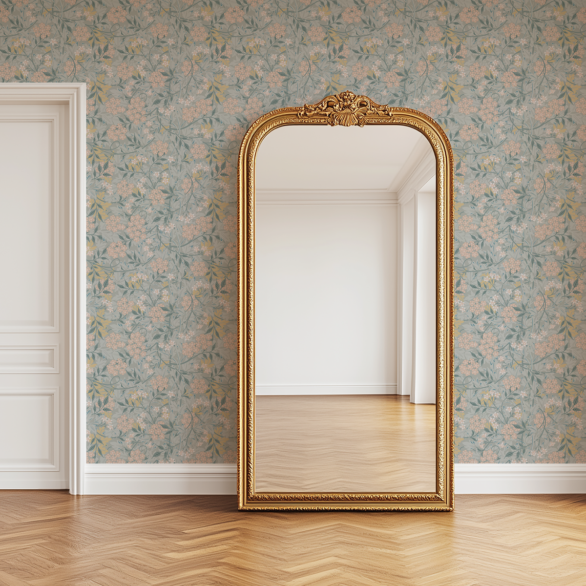An ornate gold-framed floor mirror with a baroque-style carved top, standing in a room with pale blue floral wallpaper, light wood herringbone floors, and a white door on the left.