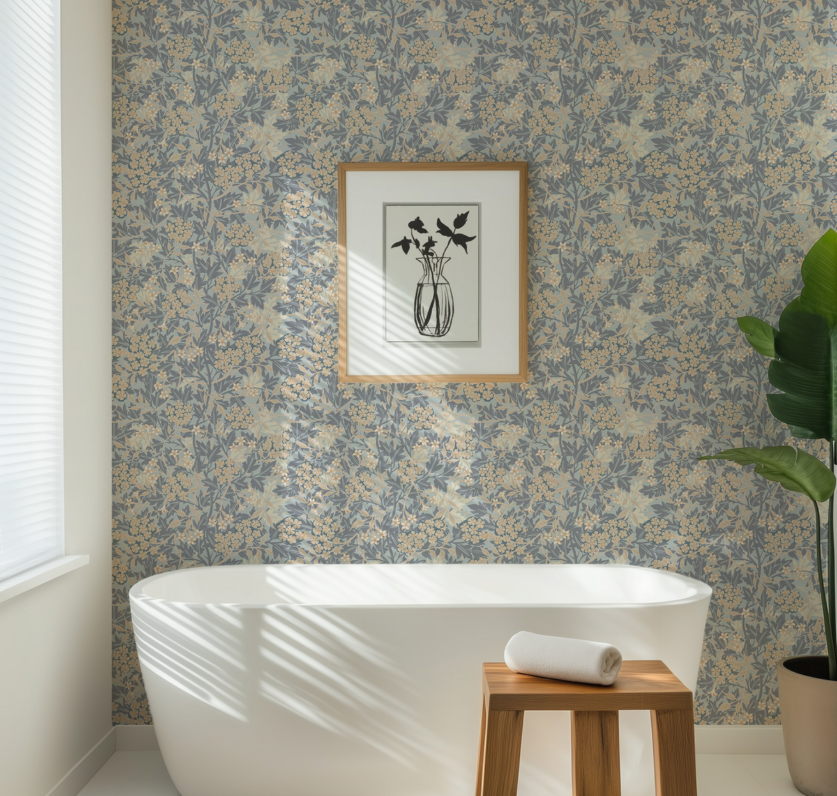 A bathroom interior featuring a blue and beige floral toile de Jouy wallpaper pattern, a white freestanding bathtub, a small wooden stool with a rolled white towel, a framed black-and-white floral print on the wall, and a potted green plant.
