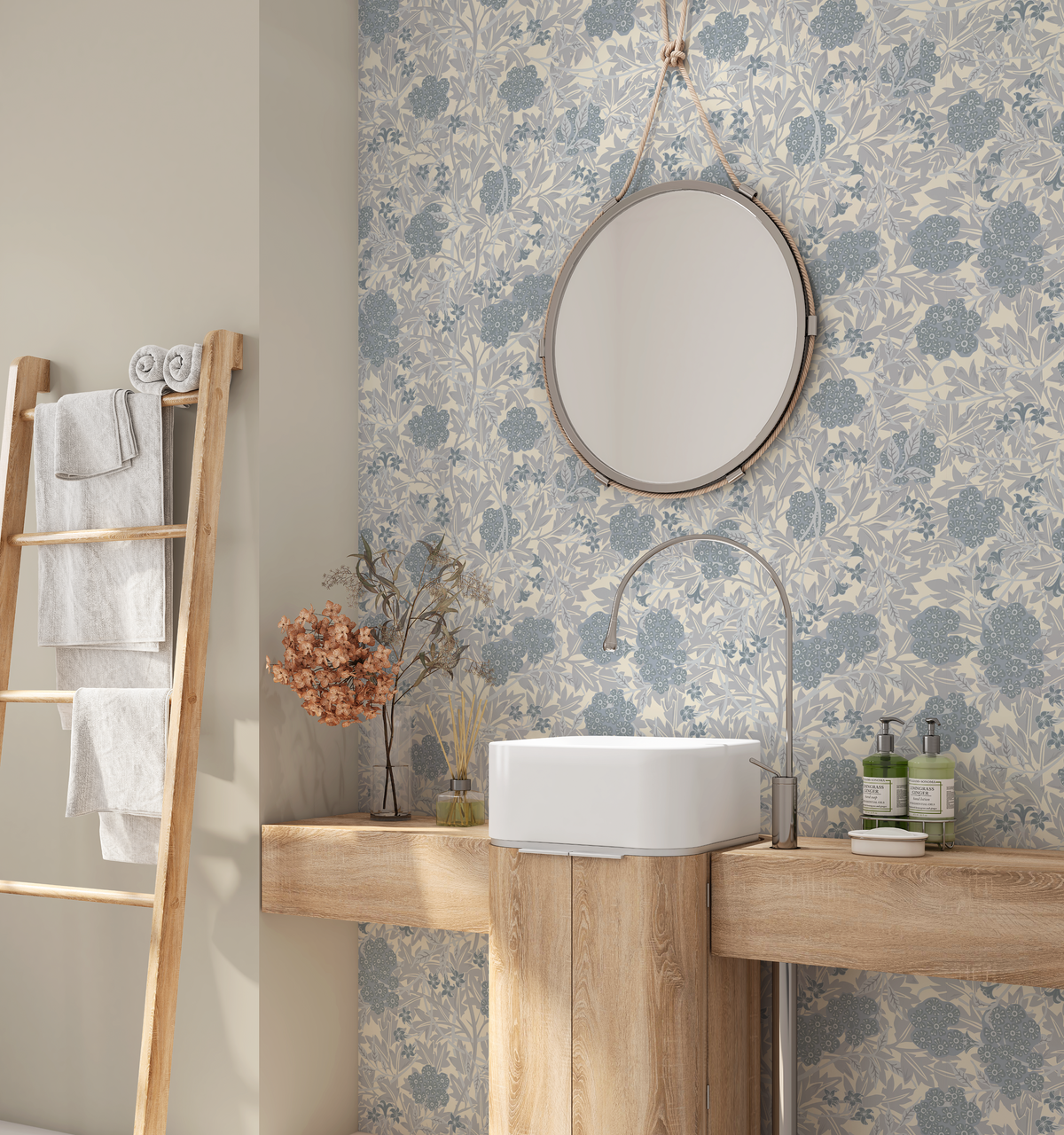 A bathroom interior featuring a light blue and gray floral wallpaper pattern, a round silver-framed mirror hanging on the wall, a white vessel sink on a wooden pedestal, and a wooden towel ladder with white towels.
