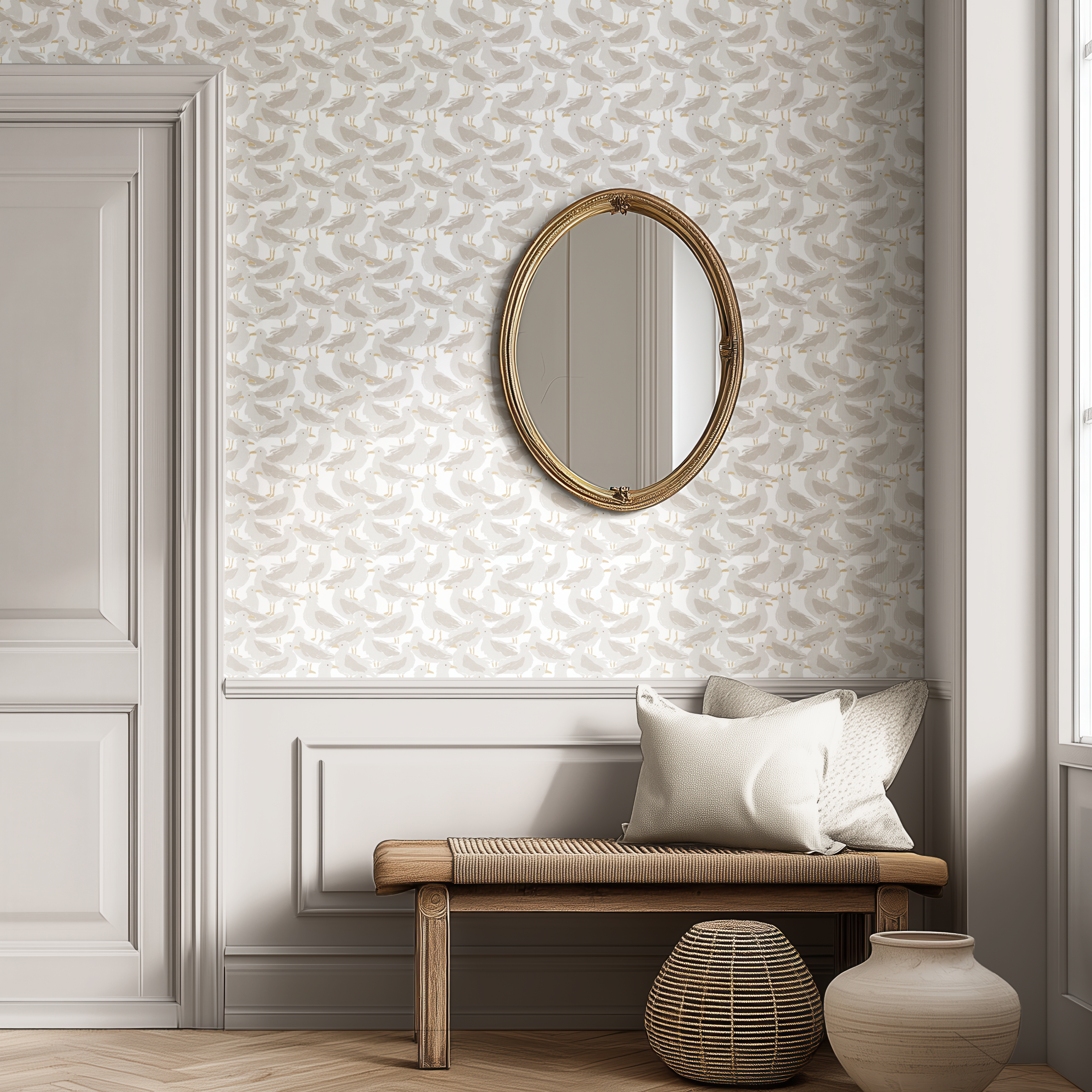 A styled interior corner with textured white wallpaper, a gold-framed oval mirror, a wooden bench with white pillows, a woven side table, and a ceramic vase.