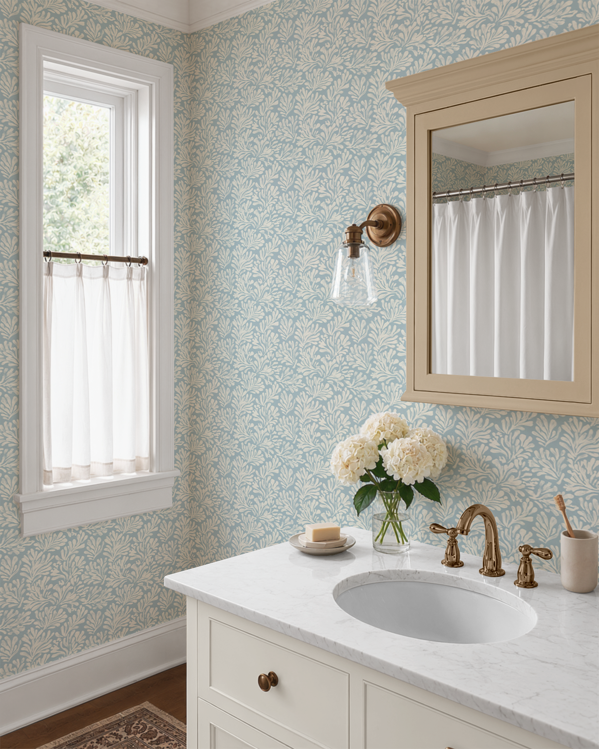 A styled bathroom interior featuring light blue and white floral wallpaper, a white vanity with a marble countertop, brass faucet hardware, a wall-mounted mirror with a light wood frame, and a window with sheer curtains.