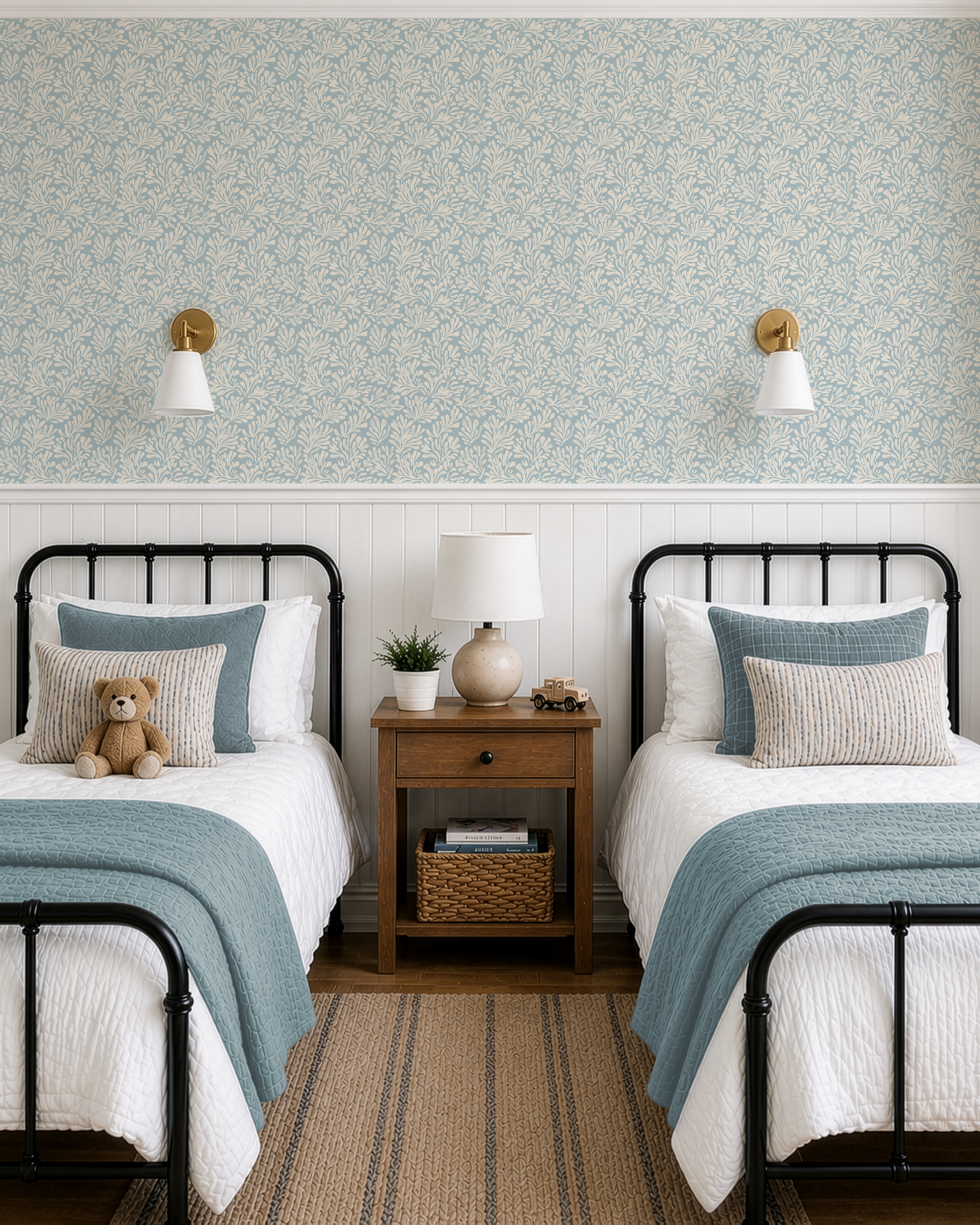 A styled children's bedroom featuring two twin beds with white bedding and teal-blue throws, flanking a wooden nightstand. The wall behind is decorated with a light blue and white floral damask wallpaper and a white wainscoting panel, accented by two gold and white wall sconces.