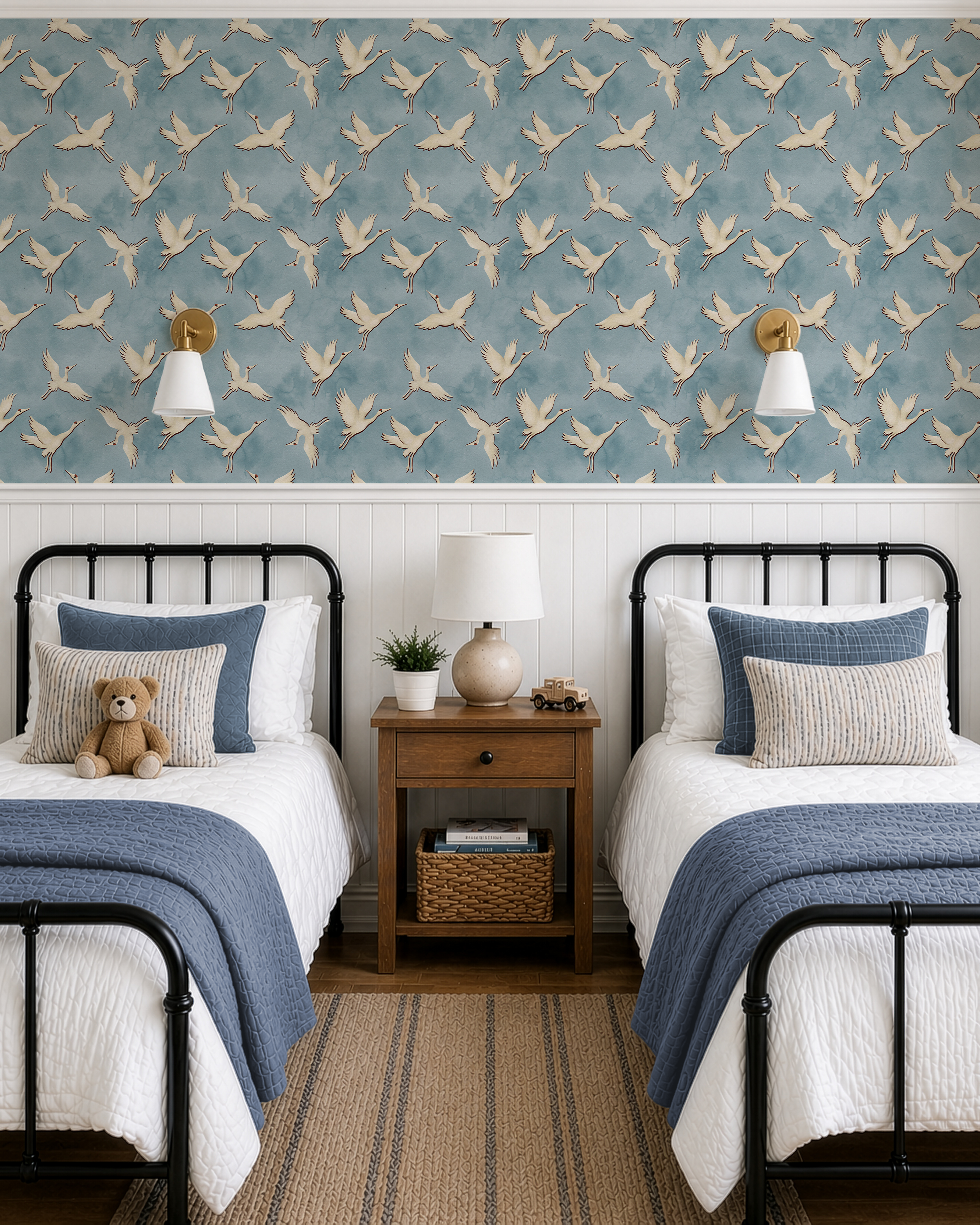 A styled children's bedroom featuring two single beds with white bedding and blue throws, set against a wall with blue wallpaper patterned with flying beige birds and white wainscoting. A wooden nightstand with a lamp and basket sits between the beds.