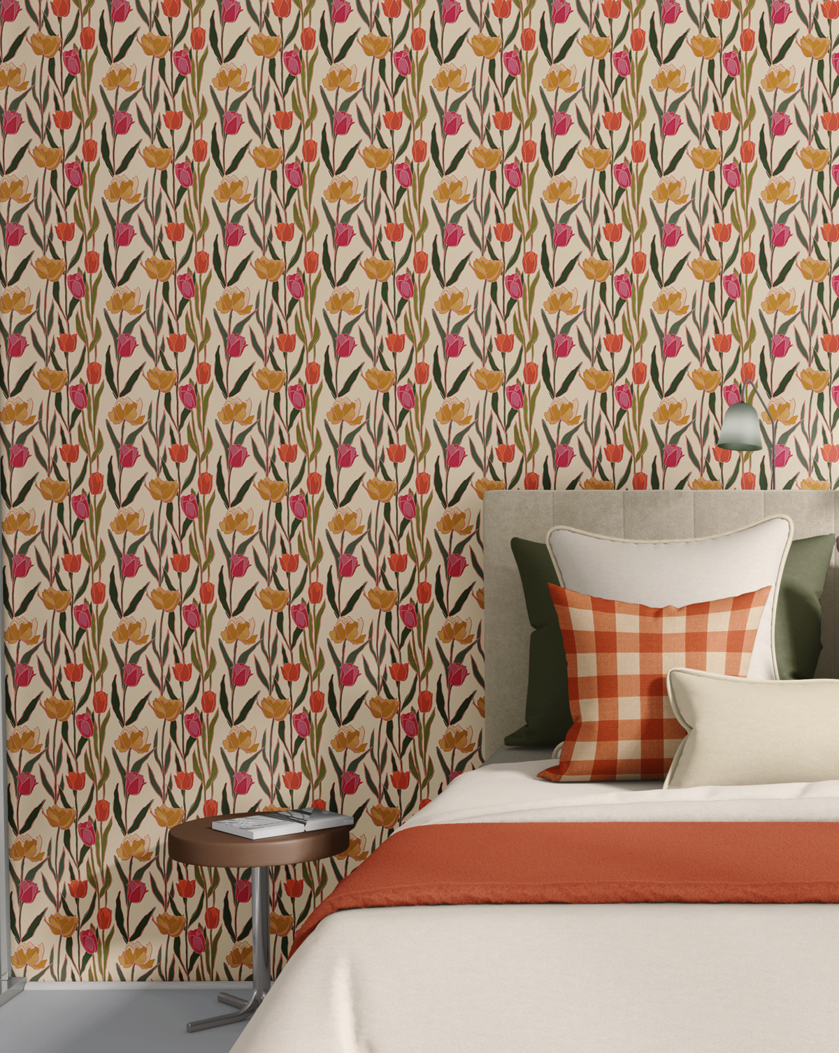 A styled bedroom scene featuring a wall covered in a dense floral wallpaper pattern with tulips in pink, orange, and yellow on a beige background. In front of the wall is a bed with white bedding, a rust-orange throw blanket, and decorative pillows, including a gingham accent pillow.