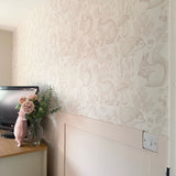 A serene nursery corner featuring wallpaper with a woodland animal theme in beige, showing rabbits, squirrels, and hedgehogs, complemented by a pale pink pig-shaped pillow and fresh flowers.