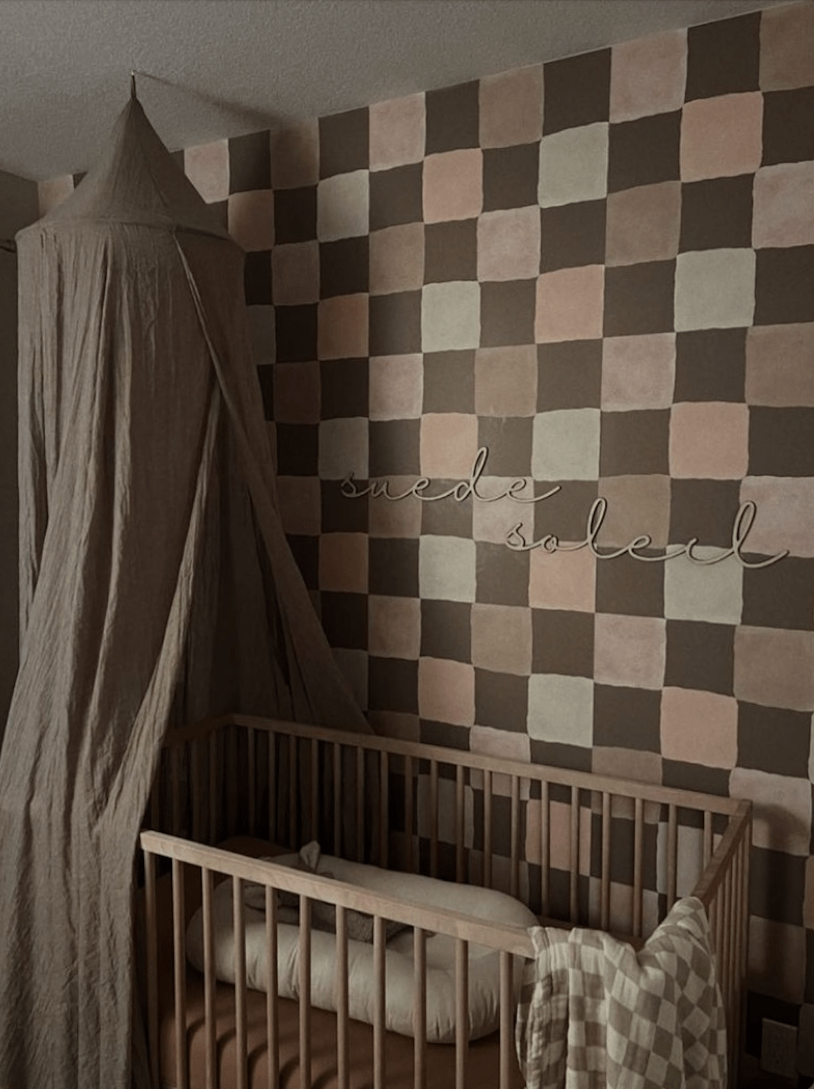 Stylish boho baby nursery featuring a checkered wallpaper in brown and blush pink tones, with a wooden crib, soft bedding, and a canopy, creating a cozy and elegant space