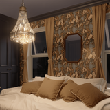 A classic bedroom interior featuring an opulent crystal chandelier and vintage floral wallpaper. The decor includes a plush bed with cream linens and matching curtains, all enhanced by rich, dark wood furniture and an elegant octagonal mirror.
