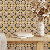 A rattan bench adorned with books and pampas grass in a decorative vase, set against a mustard yellow wallpaper featuring scalloped dusty pink medallion florals for a warm and inviting look.