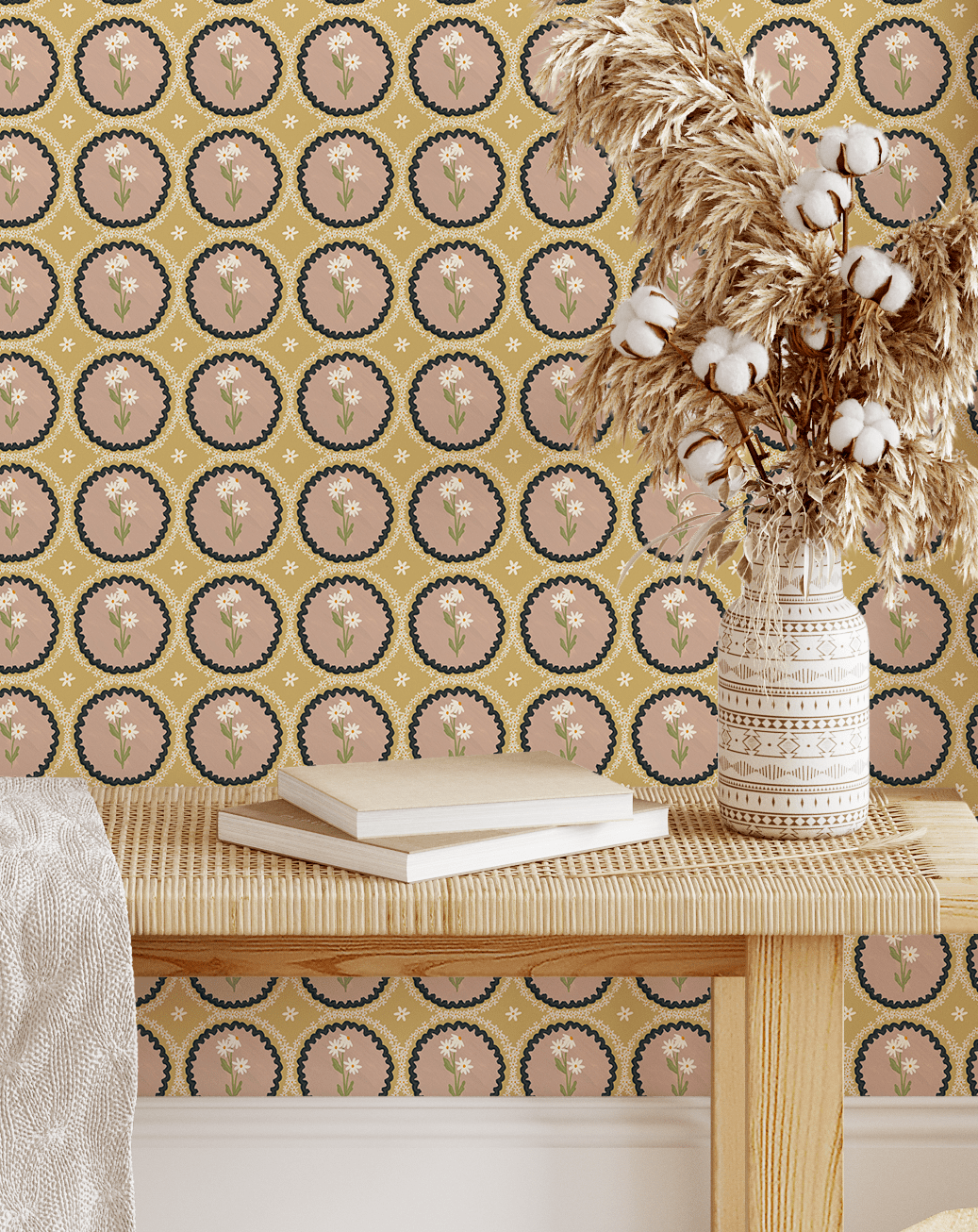 A rattan bench adorned with books and pampas grass in a decorative vase, set against a mustard yellow wallpaper featuring scalloped dusty pink medallion florals for a warm and inviting look.