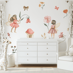 A charming nursery with fairy garden wall decals, showcasing fairies, flowers, and toadstools. The room features a white dresser, a cozy armchair, and soft white curtains, perfect for creating a serene and playful environment for a child.