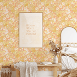 A cozy bedroom scene showcasing the Happy Floral peel and stick wallpaper on the walls. The room includes a wooden bench with a throw blanket, a woven basket, a bed with neutral bedding, and a framed picture that reads "Bloom Where You Are Planted