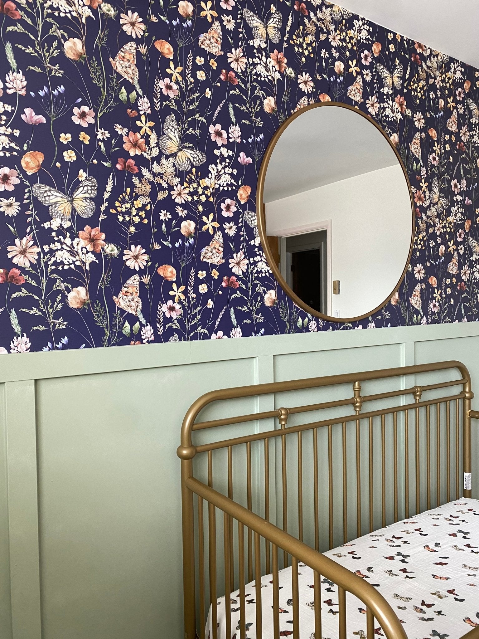 A charming nursery featuring a navy blue wallpaper adorned with detailed floral and butterfly patterns, complemented by a circular mirror and a green half-wall.