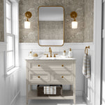 A stylish bathroom with walls covered in a neutral-toned butterfly and floral wallpaper. The decor includes a white vanity with gold handles, a white marble top, and brass fixtures.