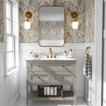 A well-lit bathroom interior with walls covered in a cream leaf pattern wallpaper that matches the previous image, completed with a wooden vanity with gold accents and a rectangular mirror.