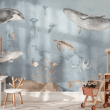 A children's playroom featuring an ocean-themed wall mural. The mural showcases a variety of sea creatures including orcas, sharks, whales, jellyfish, sea turtles, and an octopus, all painted in a soft watercolor style. The room is furnished with a small white table, wooden chairs, and a toy cart filled with plush toys and children's books, enhancing the marine nursery decor.