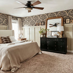  A cozy bedroom featuring an olive branch wallpaper border, beige walls, a large bed with beige bedding, and a dark wood ceiling fan. A black dresser with a gold-framed mirror adds a classic touch.