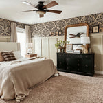 A cozy bedroom featuring an olive branch wallpaper border, beige walls, a large bed with beige bedding, and a dark wood ceiling fan. A black dresser with a gold-framed mirror adds a classic touch.