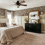 A cozy bedroom featuring an olive branch wallpaper border, beige walls, a large bed with beige bedding, and a dark wood ceiling fan. A black dresser with a gold-framed mirror adds a classic touch.