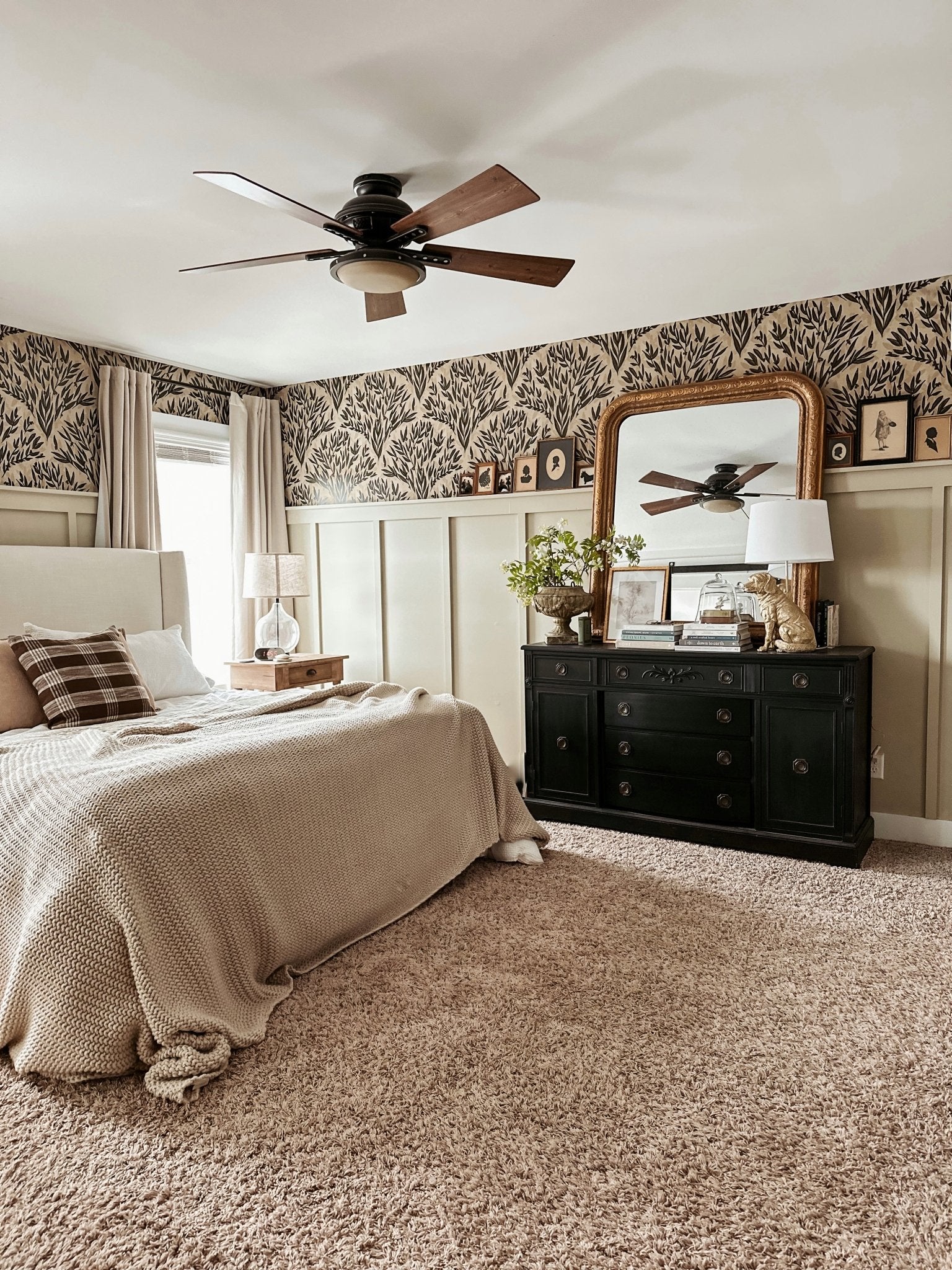 A cozy bedroom featuring an olive branch wallpaper border, beige walls, a large bed with beige bedding, and a dark wood ceiling fan. A black dresser with a gold-framed mirror adds a classic touch.
