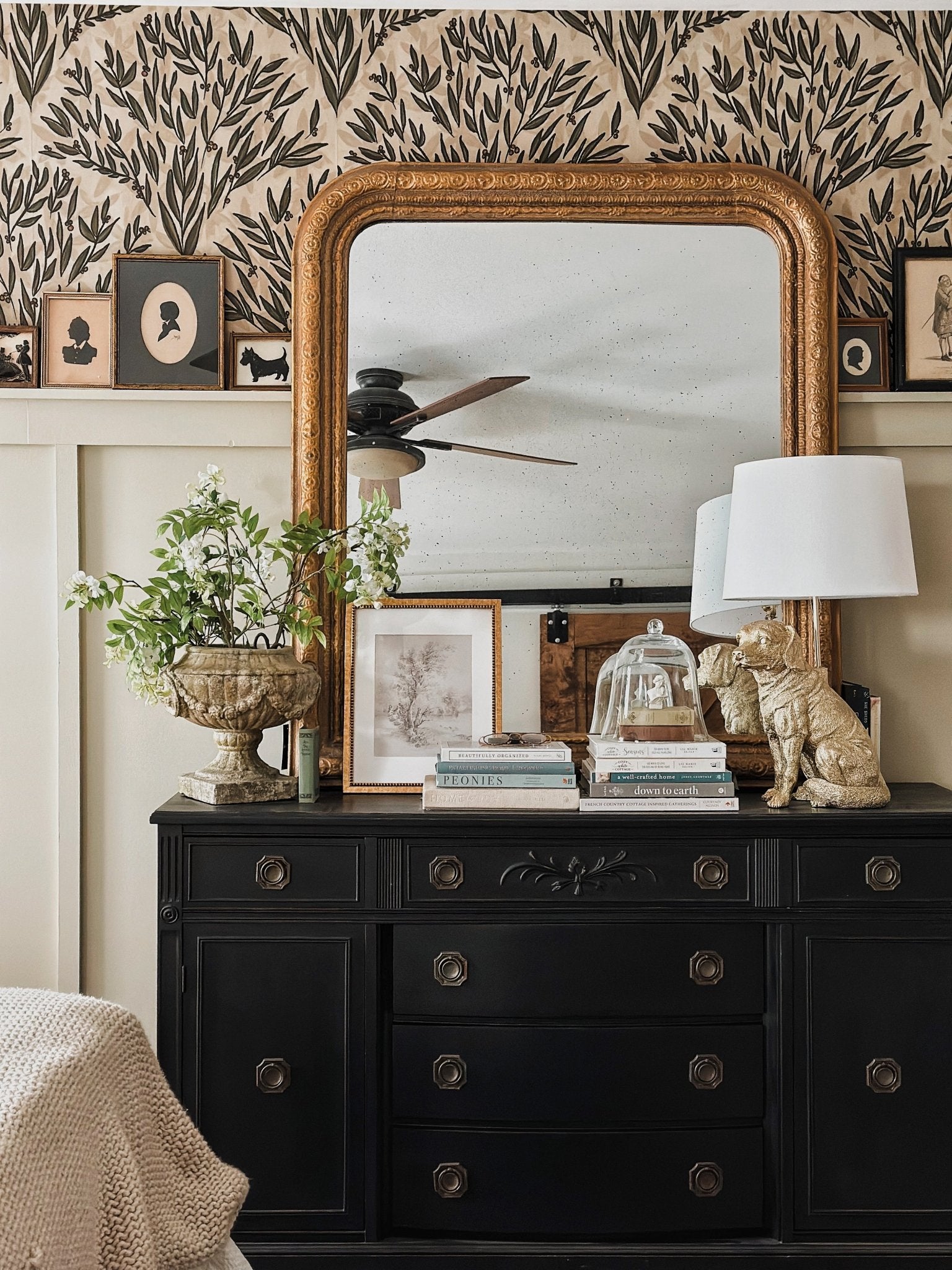 Close-up of the bedroom's decor, highlighting the black dresser adorned with books, plants, and framed pictures against the decorative olive branch wallpaper.