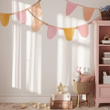 A playroom with a pastel bunting banner wall decal in pink, yellow, and orange. The decal hangs above a pink bookcase filled with toys and decor, including a stuffed rabbit and wicker basket, perfect for a playful, pastel-themed space.
