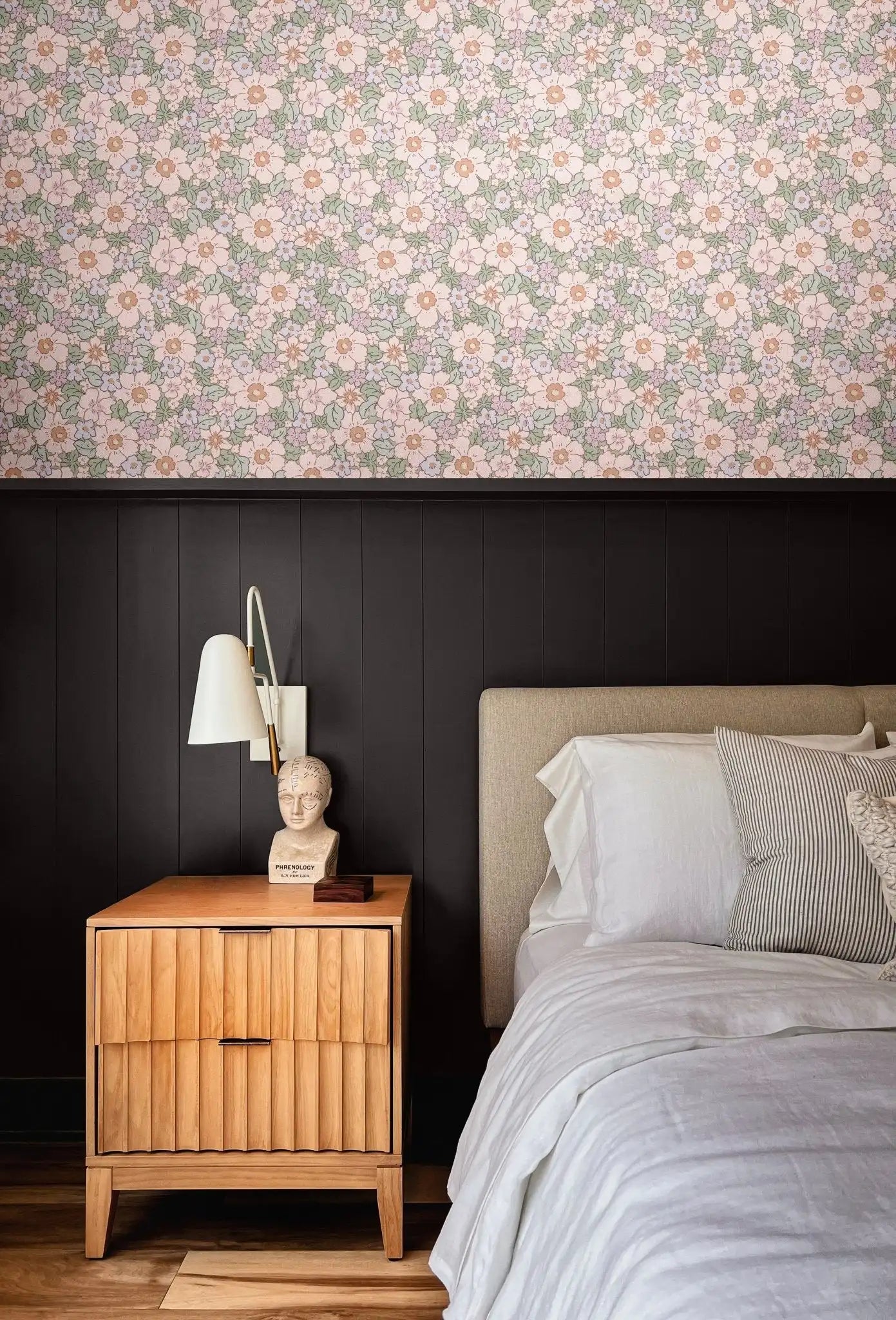 Romantic floral wallpaper above black wainscoting in cozy modern bedroom with wood nightstand and neutral bedding