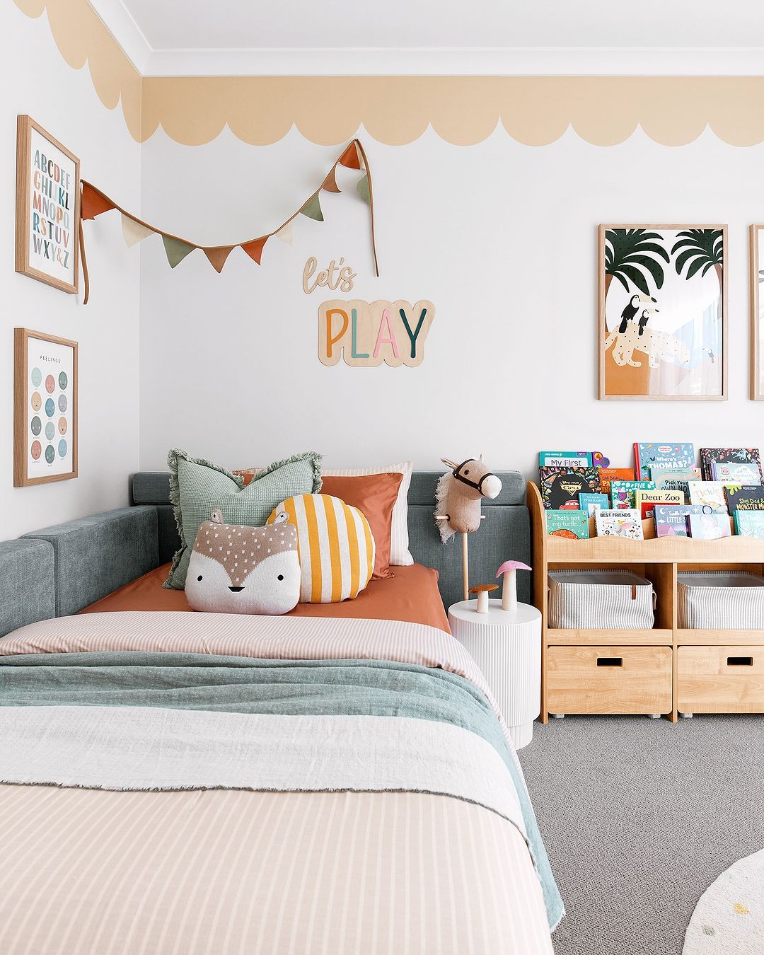 A children's bedroom showcasing a bed with colorful bedding and cushions, a wall-mounted bookshelf filled with books, and framed artwork. A scalloped wallpaper border in a soft peach hue accents the top of the walls, creating a playful atmosphere