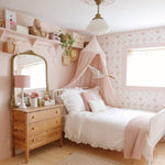 A cozy, vintage-style girl's bedroom with floral wallpaper, a white bed with pink canopy, wooden dresser, and decorative items on shelves. The room has a warm, inviting feel with soft pink and white tones.