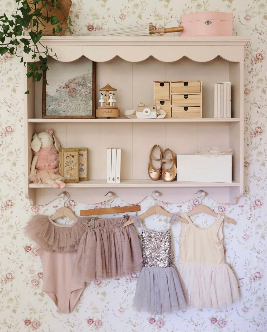 A vintage-inspired children's room with floral wallpaper, a pink wall shelf, and assorted decorative items. The shelf holds a framed painting, books, wooden storage boxes, and ballet shoes, with delicate dresses hanging below.