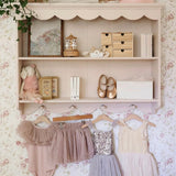 A vintage-inspired children's room with floral wallpaper, a pink wall shelf, and assorted decorative items. The shelf holds a framed painting, books, wooden storage boxes, and ballet shoes, with delicate dresses hanging below.