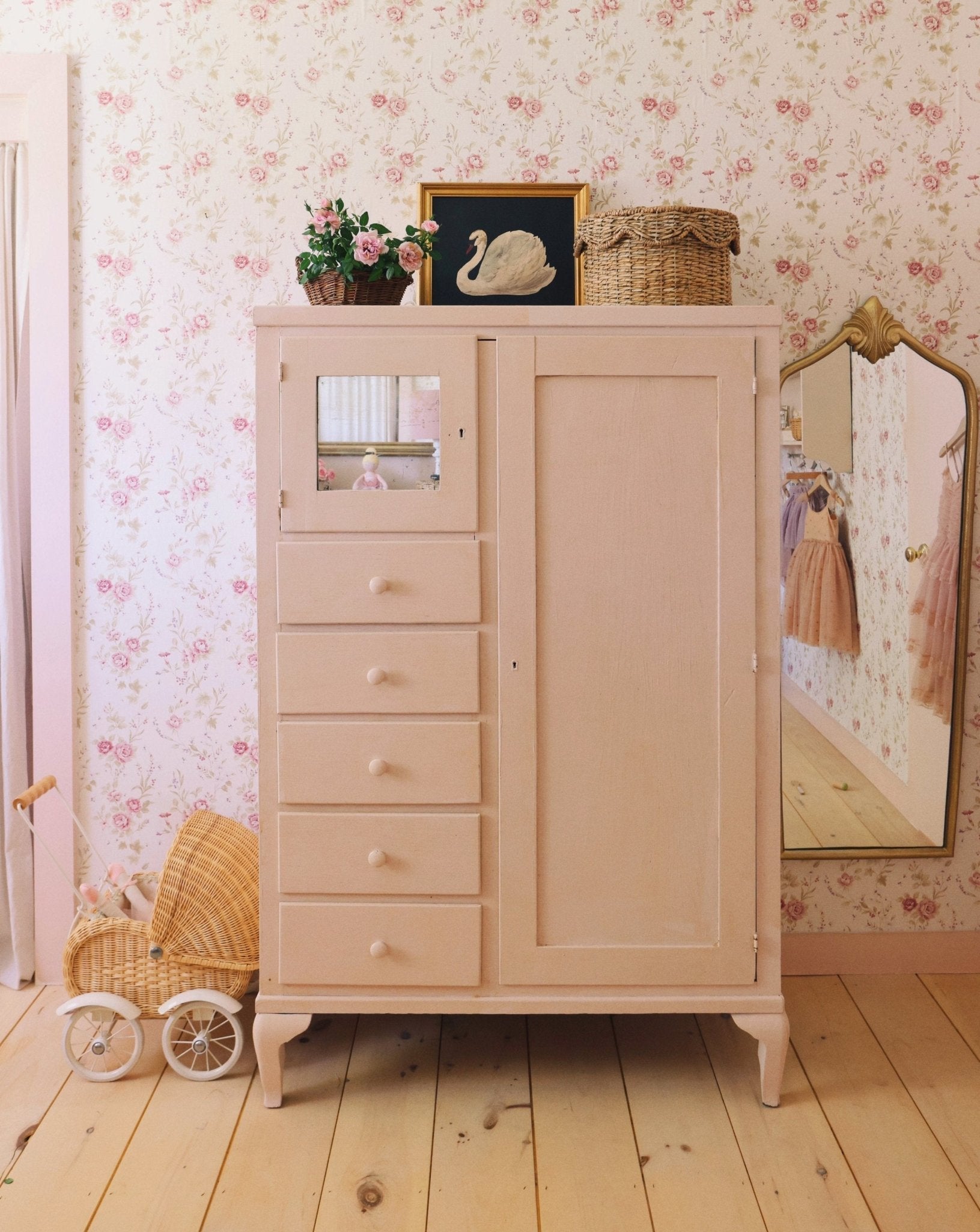 A vintage-inspired room with a pink painted dresser, wicker basket, and floral wallpaper. The dresser is decorated with a framed swan painting and pink flowers, creating a charming, antique aesthetic.