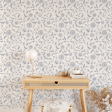 A cozy workspace with blue floral self-adhesive, removable wallpaper. A stylish wooden desk with angled legs holds a lamp, books, and small gold accessories. A fluffy stool and a vase with dried plants add warmth to the space