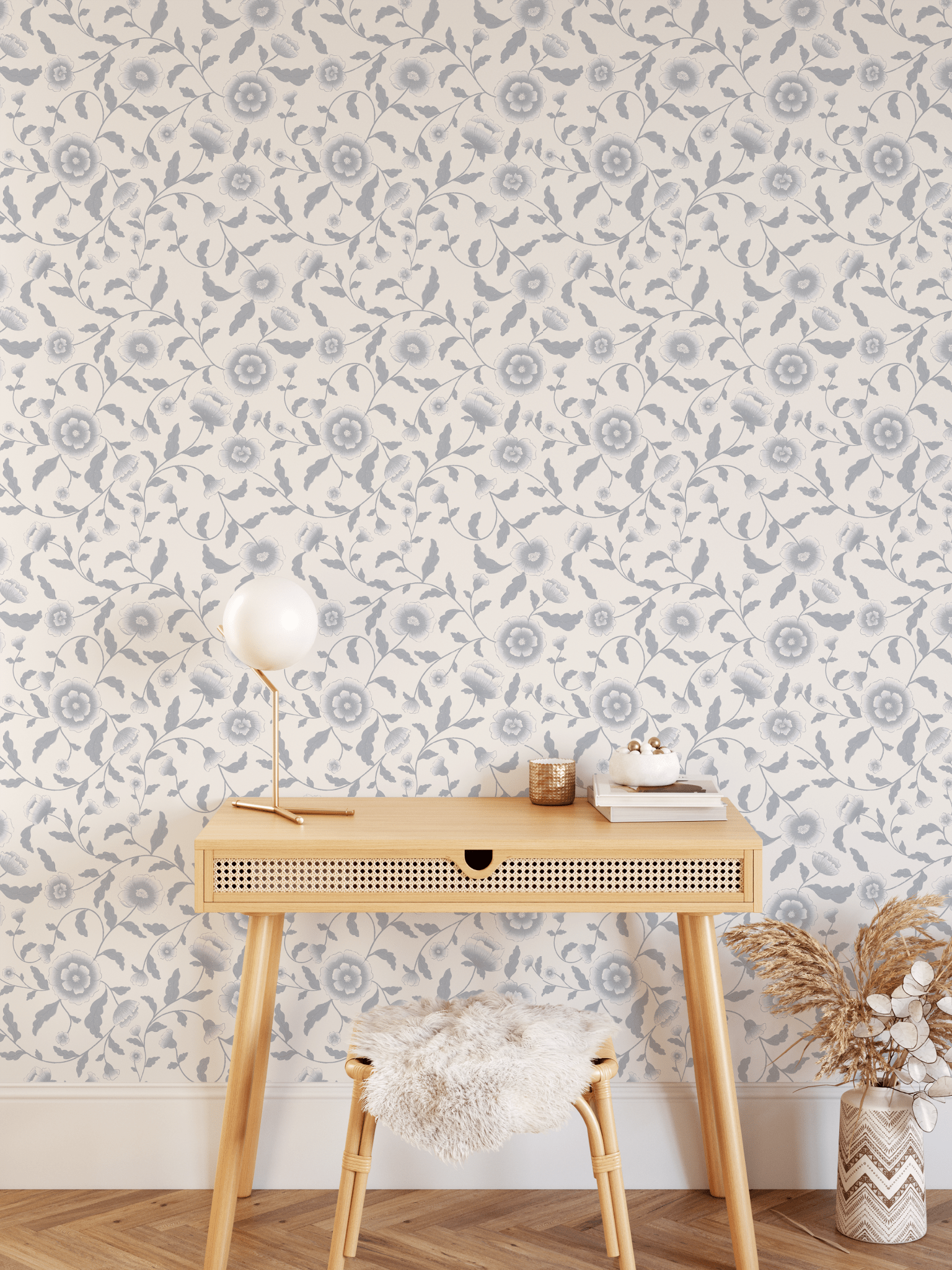 A cozy workspace with blue floral self-adhesive, removable wallpaper. A stylish wooden desk with angled legs holds a lamp, books, and small gold accessories. A fluffy stool and a vase with dried plants add warmth to the space