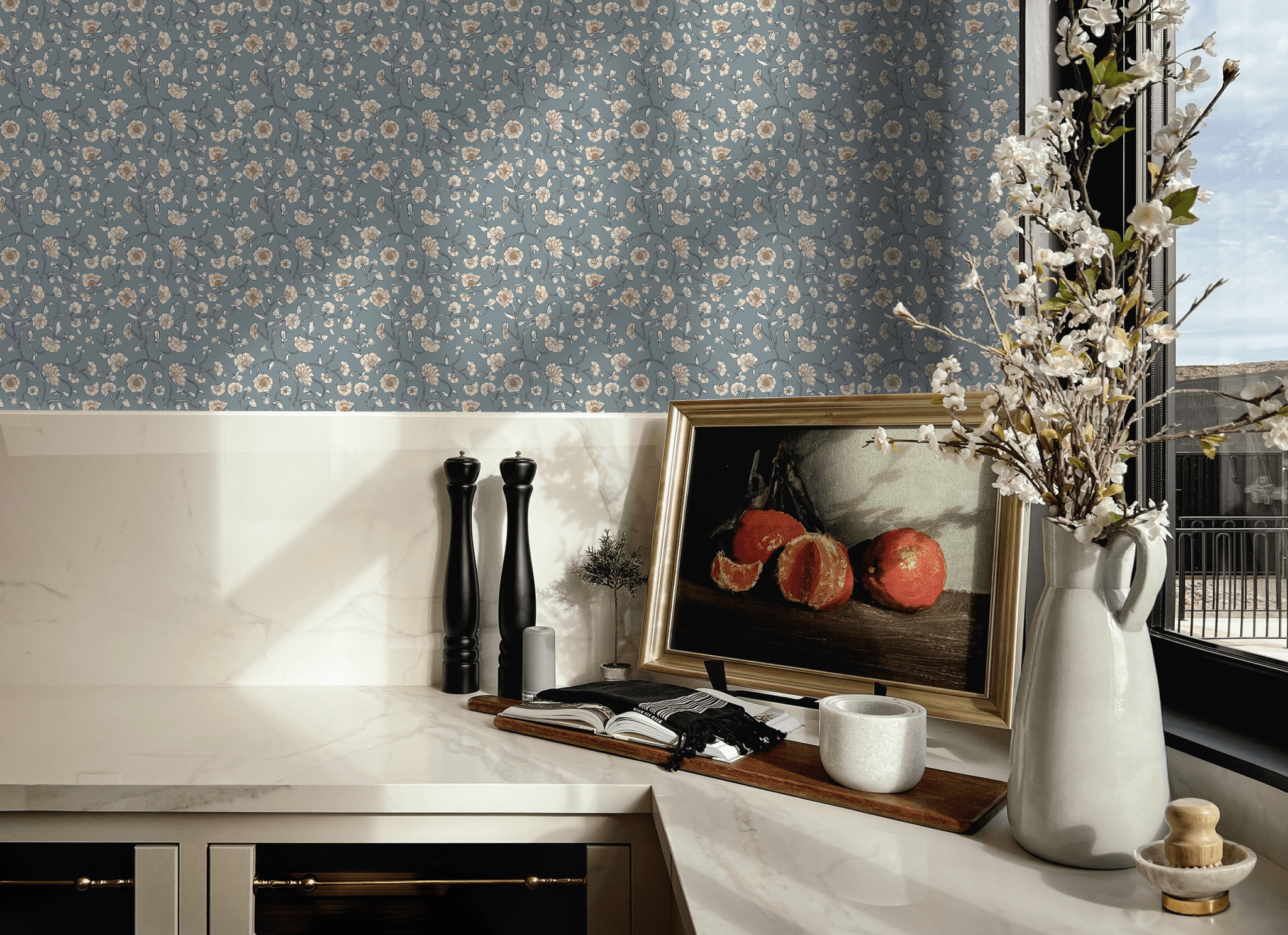 A stylish kitchen corner with blue vintage floral wallpaper. The countertop displays elegant salt and pepper mills, a framed picture of pomegranates, a ceramic pitcher with blossoming branches, and a modern candle holder