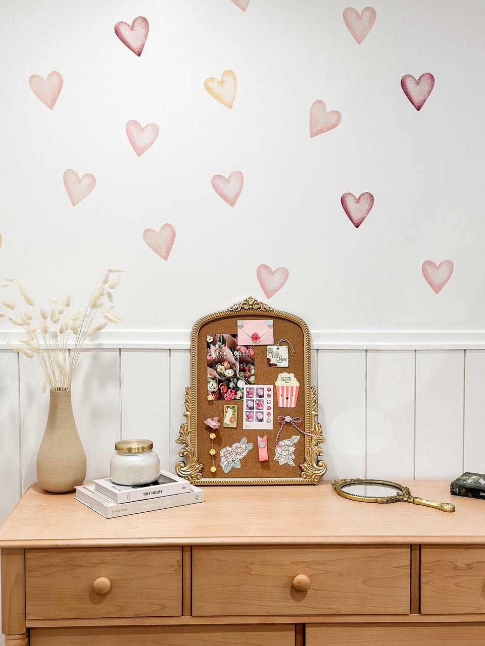 A charming boho-style dresser with a vintage gold-framed corkboard decorated with stickers and notes, flanked by a vase of dried flowers, books, a candle, and a handheld mirror.