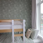 Pine bunk bed with white accents in a room with forest green floral wallpaper, adjacent to a window with sheer white curtains.