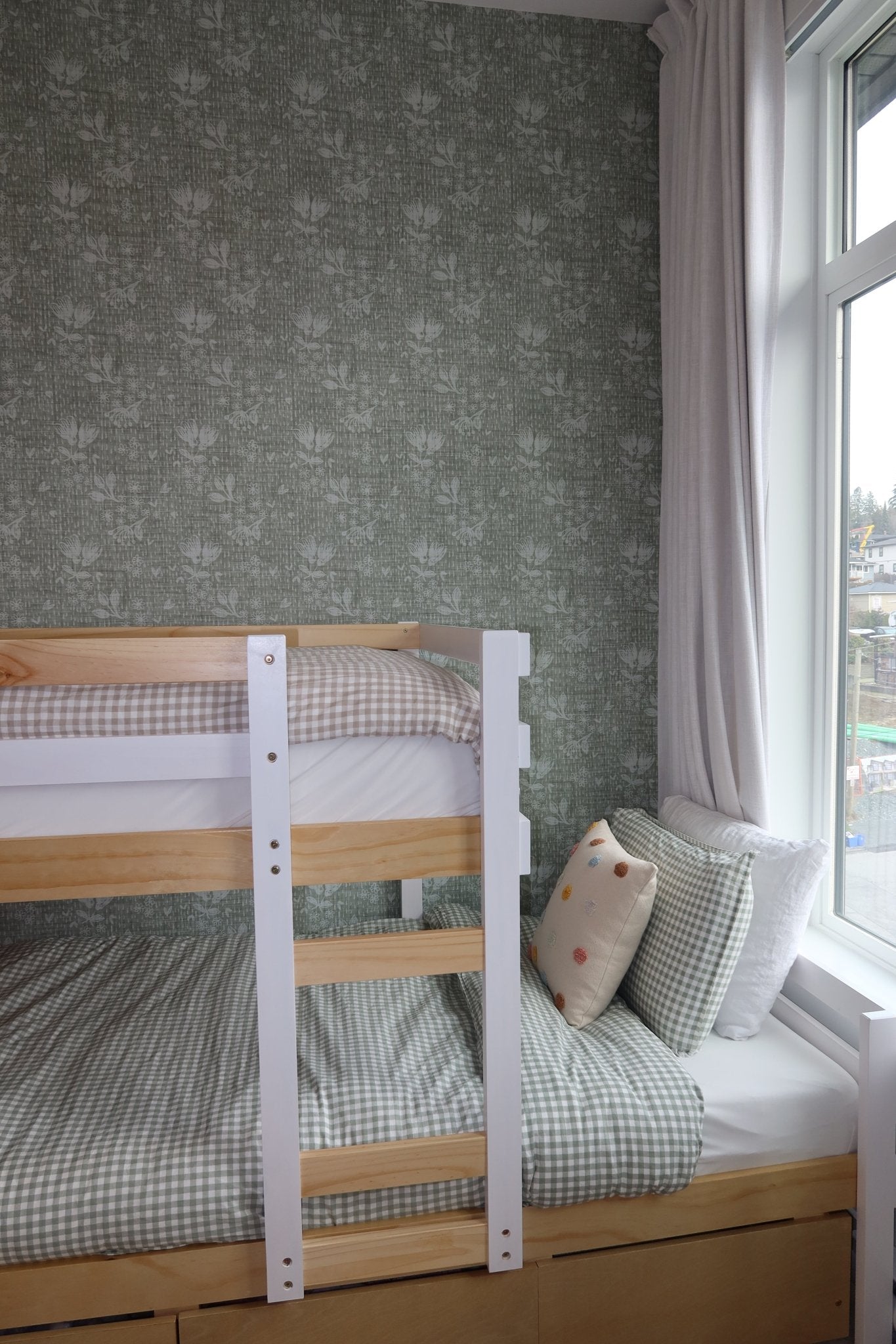 Pine bunk bed with white accents in a room with forest green floral wallpaper, adjacent to a window with sheer white curtains.