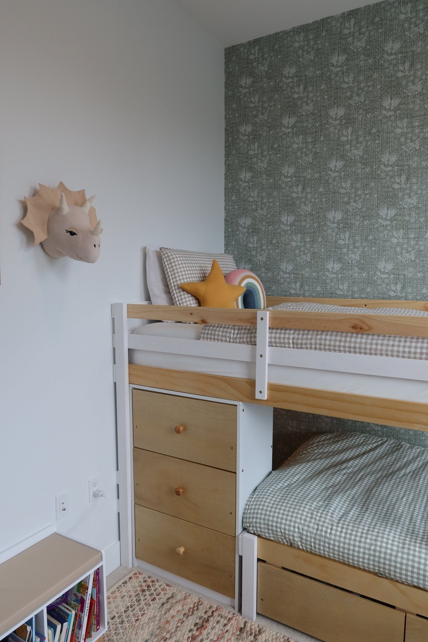 A cozy corner of a child's bedroom with a pine bunk bed, forest green floral wallpaper, and assorted plush toys and cushions.