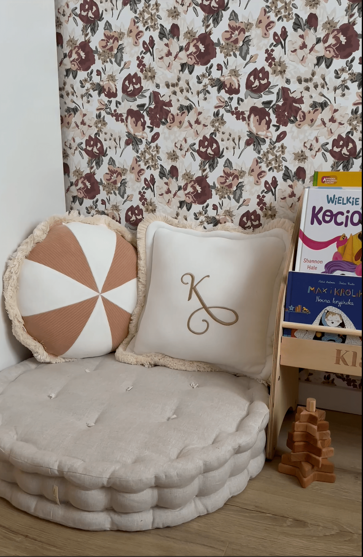A cozy reading nook with a large cushioned floor seat, decorative pillows, and a floral wallpaper featuring maroon and taupe flowers