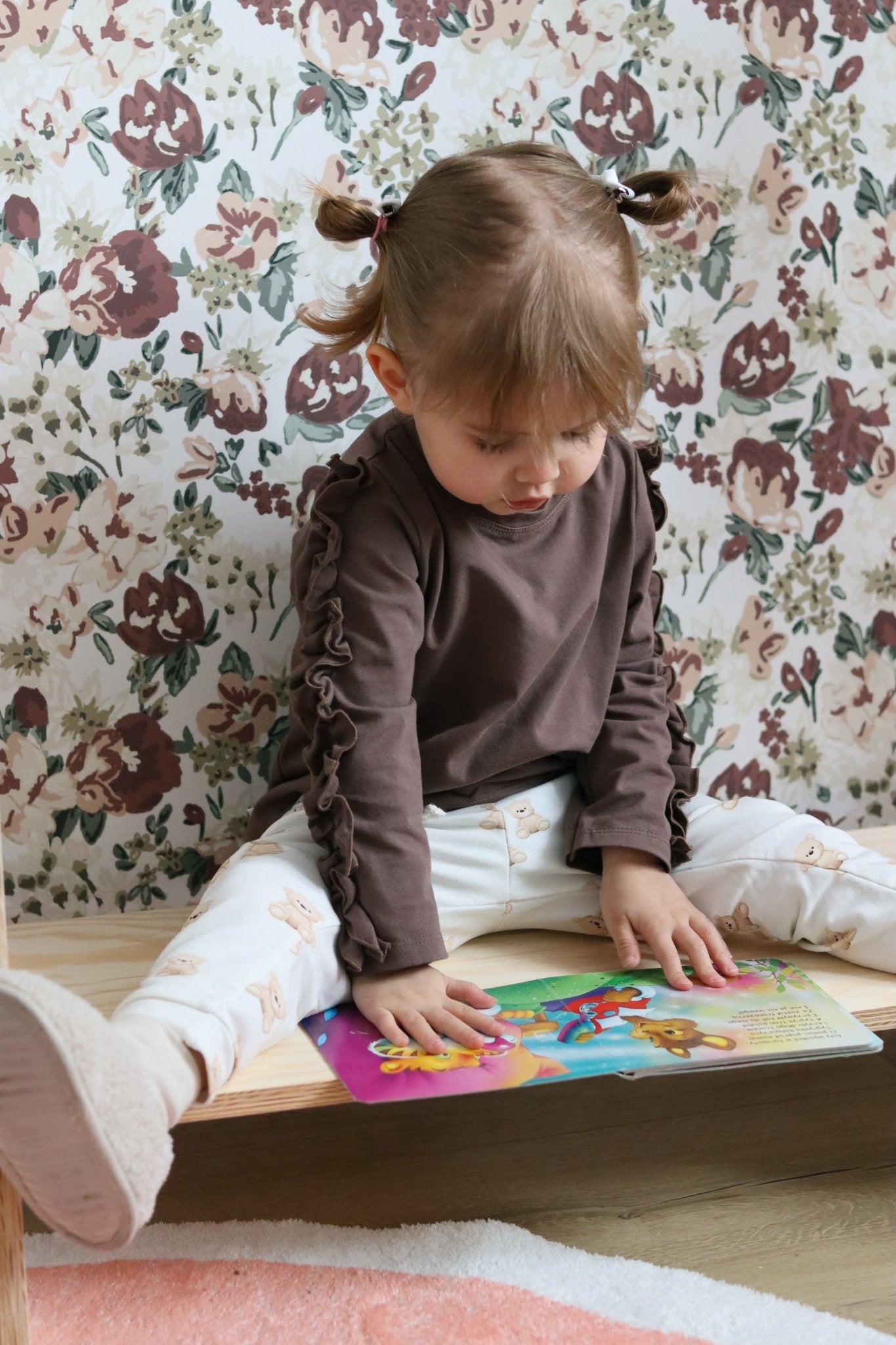 A young child sits on a cushioned floor seat, reading a book against a backdrop of floral wallpaper with maroon and olive heirloom-style flowers