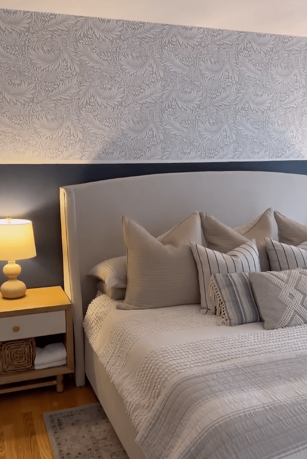 Cozy bedroom with whimsical grey and white leaf-patterned wallpaper, a neatly made bed with textured pillows, wooden nightstand, and ceramic table lamp.