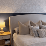 Cozy bedroom with whimsical grey and white leaf-patterned wallpaper, a neatly made bed with textured pillows, wooden nightstand, and ceramic table lamp.