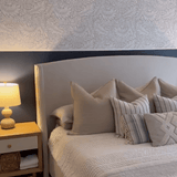 Cozy bedroom with whimsical grey and white leaf-patterned wallpaper, a neatly made bed with textured pillows, wooden nightstand, and ceramic table lamp.