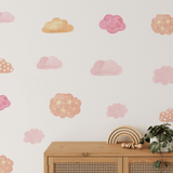 A nursery room wall decorated with watercolor wall stickers of clouds in shades of pink and gold, with a wooden toy rainbow on a cabinet and a hanging plant