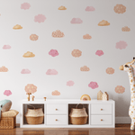 A child's room with a white wall adorned with pink and orange pastel cloud decals, a stuffed blue whale on a wicker basket, and a tall giraffe toy next to a white storage unit.