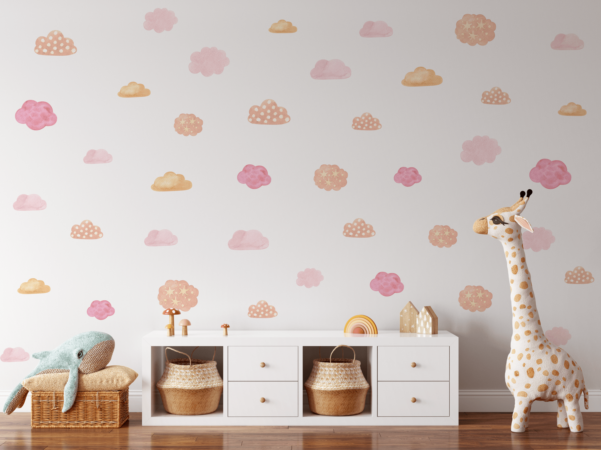 A child's room with a white wall adorned with pink and orange pastel cloud decals, a stuffed blue whale on a wicker basket, and a tall giraffe toy next to a white storage unit.